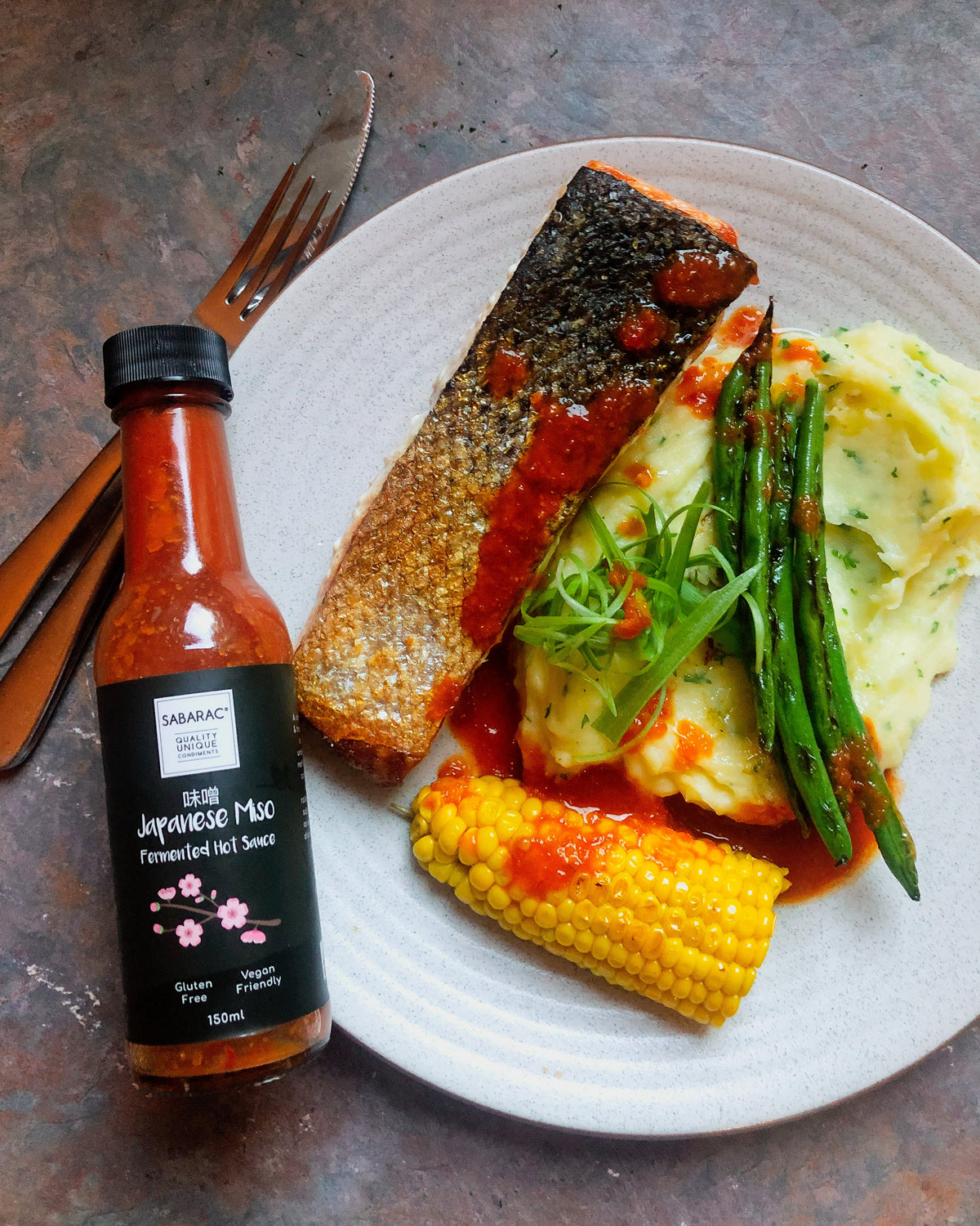 A bottle of Sabamac Japanese Miso Forwarded Hot Sauce placed next to a plated meal featuring salmon, mashed potatoes, green beans, and corn on a rustic dark gray surface.